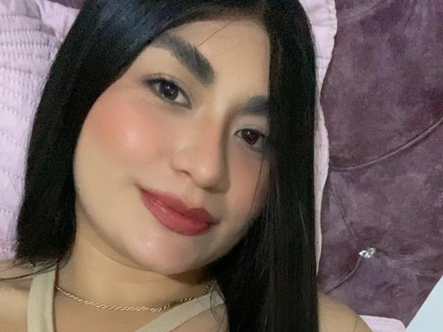 MiaDuboisX with black hair, light skin, and red lipstick, wearing a gold necklace, smiling at the camera, showing her slightly chubby face and soft, shiny skin.