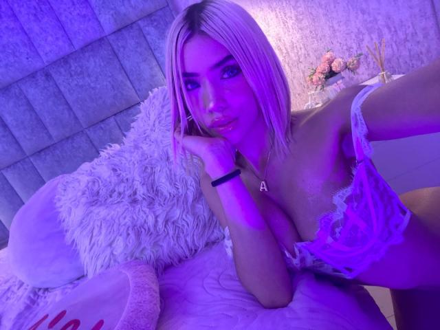ChristieGarci with blonde hair, purple lighting, wearing white lace lingerie, resting head on hand, showing large tits, pink lipstick, white fluffy pillow, and floral vase in background.