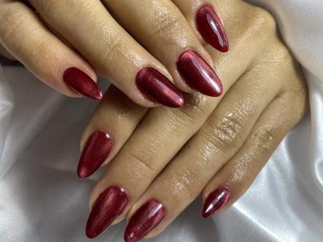 MiaDuboisX's hands with shiny, metallic red, almond-shaped nails, resting on a white satin surface. Her skin is smooth, and the nails are glossy and well-manicured.
