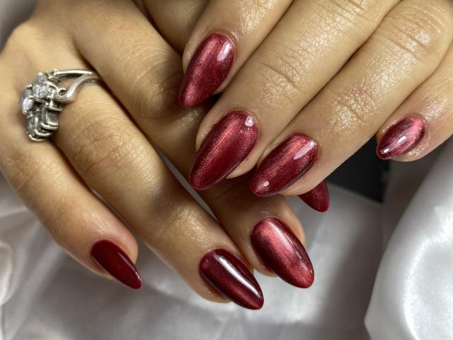 Close-up of MiaDuboisX's hands with shiny, metallic red nails. Her fingers are slightly parted, showing off the long, oval-shaped nails with a smooth, reflective texture. A silver ring with a cluster of small, clear diamonds is on her left ring finger. The skin tone is light brown, and the nails have a glossy finish. The white fabric in the background contrasts with the dark red nails. The image highlights the elegance and detail of the manicure.