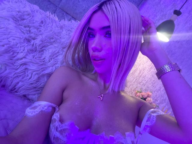 ChristieGarci with blonde bob, purple lighting, white lace bra, showing tits, hand in hair, silver watch, necklace, lying on fluffy pillow, looking at camera, subtle smile.