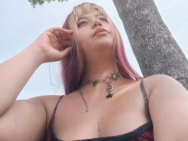 BelleVivienneHt hat pinke und blonde Haare, trägt eine schwarze und rote Spitzenbluse, die ihre Brüste zeigt, zusammen mit einer Halskette, und ihre Hand ist in der Nähe ihres Gesichts, unter einem Baum..