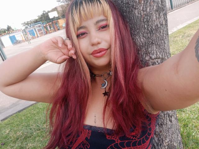 BelleVivienneHt stands in front of a tree with her right arm raised, holding her hair. She has long, red-to-blonde ombre hair with straight bangs. She wears a black, red-patterned top and a black choker with a crescent moon and star. Her makeup includes pink eyeshadow and red lipstick. Her skin is light brown. The grass and background are blurred, focusing on her. She has a small tattoo on her right arm. Her expression is relaxed.