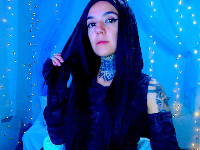 LannieGonzales tiene el pelo largo y negro, un cuello tatuado y una blusa de encaje negro. En el fondo hay luces azules y pequeñas luces de hadas. Ella está mirando a la cámara..