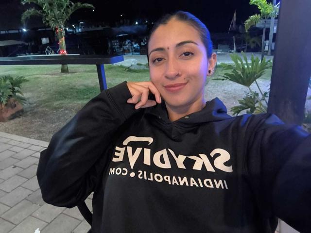 ZaraBeicker, with dark hair in a ponytail, wearing a black Evolv hoodie, smiling slightly, resting chin on hand, showing small nose and earrings, in a night-time outdoor setting.