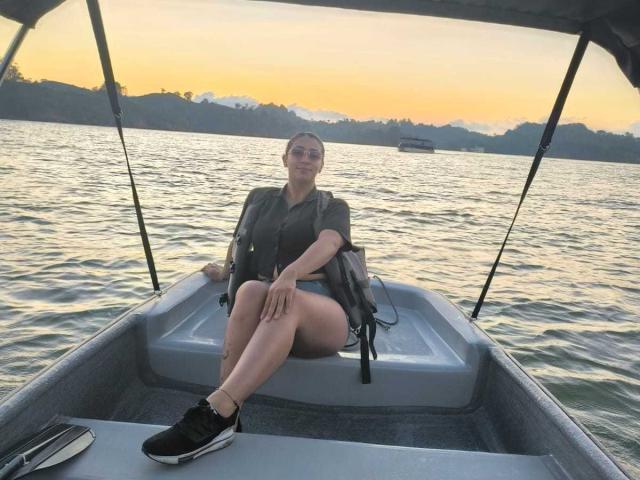 ZaraBeicker, sitting in a boat, wearing a green shirt and black shorts, with a life jacket, and black sneakers, legs crossed, smiling, sunset in the background.