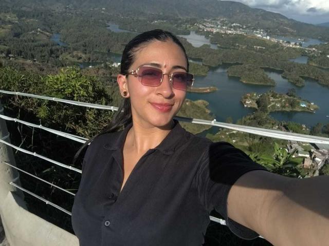 ZaraBeicker has medium tits, wearing a black shirt, pink sunglasses, and ear gauges, taking a selfie with a mountain and lake view in the background.