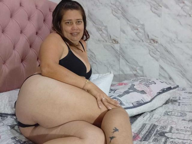ThomsonsMia, with brown hair, black bra, and black panties, sits on a bed, showing her big ass and small tits, smiling at the camera, with tattoos on her thigh.