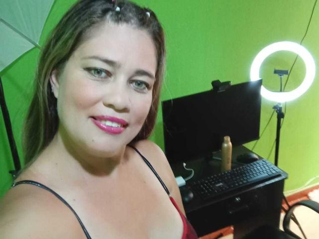 ThomsonsMia with pink lipstick, smiling, brown hair, black spaghetti straps, red top, ring light, computer, and dildo on desk. Her tits are slightly visible, she's looking at camera.