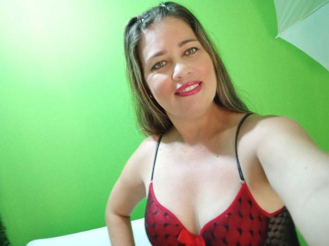 ThomsonsMia with long brown hair, red and black lace bra, smiling, showing her tits, red lipstick, green background, and white bed. She's holding the camera for a selfie.