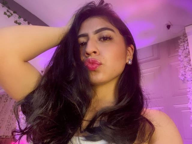 ZoeRedds with long black hair, brown skin, and pink lipstick, pouting her plump ass, wearing a white top, hand in hair, purple and pink light, earrings.