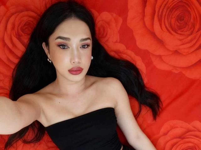 xGoddessJazzyx with black hair, wearing a strapless black top, lying on a red rose-patterned background, showing her tits, pouty red lips, and dark makeup.