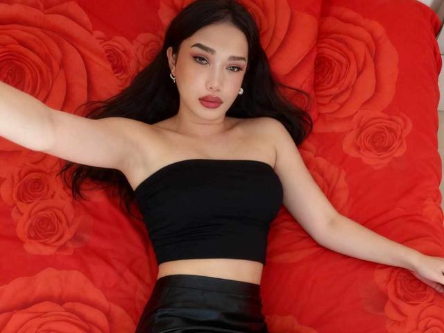 xGoddessJazzyx with long black hair, wearing a strapless black top and black leather skirt, lying on red rose-patterned bed, arms spread, looking at camera, red lipstick.