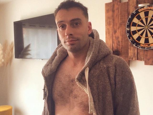 MoranS, shirtless, with a hairy chest, wearing a grey, fuzzy hoodie, has short dark hair and a slight smirk. A dartboard is on the wooden wall behind him.