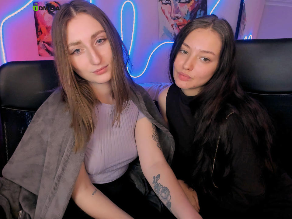 EvaYNiki with brown hair, pink shirt, gray blanket, and tattoo on left arm, sitting next to EvaYNiki with black hair, black shirt, pouting. Neon lights in background.