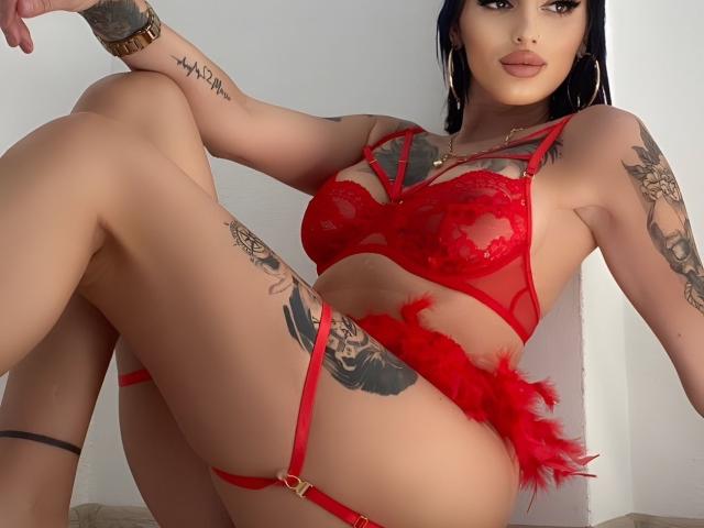 MelissaBc with red lace lingerie, tattoos on arms and thighs, black hair, hoop earrings, sitting with legs crossed, showing her tits and ass. Red garter, feathered bottom.