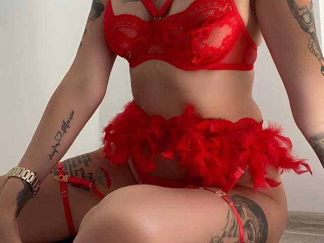 MelissaBc wearing red lace lingerie with feather accents, showing her tits and pussy in the garter. She has tattoos, a gold watch, and is sitting on a wooden floor.