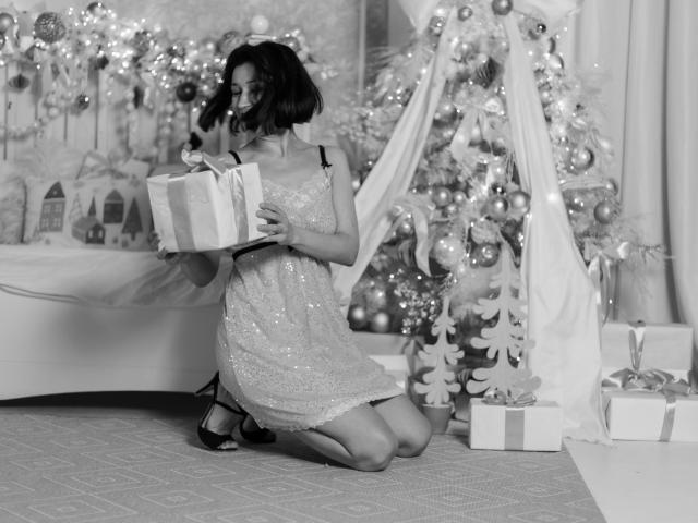 Cameron Enns está arrodillada, vestida con un vestido brillante, sosteniendo una caja de regalo y sonriendo. En el fondo se ve un árbol de Navidad y regalos. Los senos están cubiertos por el vestido..
