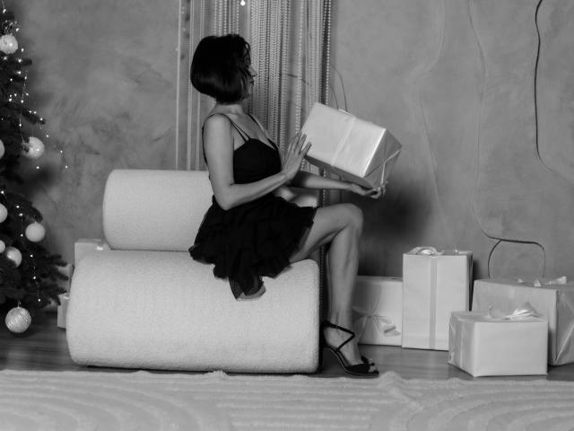 CameronEnns, sentada en una silla blanca, vestida con un vestido negro, sosteniendo una caja de regalo, mostrando sus glúteos, junto a regalos de Navidad, en una fotografía en blanco y negro..