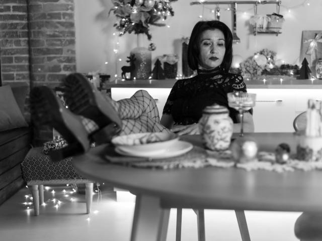 CameronEnns with dark bob, lace top, fishnet stockings, black boots, legs up, black lipstick, sitting on kitchen counter, black-and-white photo, decorative table in foreground.