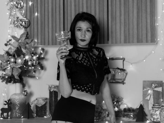 Cameron Enns mit kurzen, dunklen Haaren, einem schwarzen Spitzen-Crop-Top, einem Weinglas in der Hand, stehend, schwarze Hose, blickt in die Kamera, Weihnachtsdekoration, ein Baum mit Ornamenten, Lichterketten, schwarzer Lippenstift..