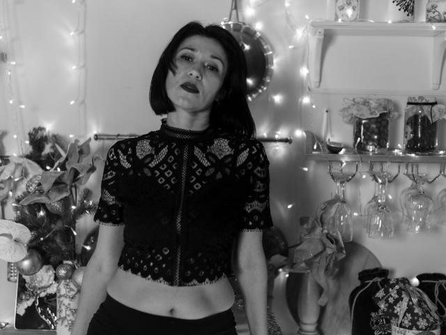 Cameron Enns, con un corte de pelo bob oscuro, vistiendo una blusa negra de encaje, mostrando su abdomen, con lápiz labial oscuro, de pie, mirando hacia arriba, rodeada de luces de hadas, en una fotografía en blanco y negro..