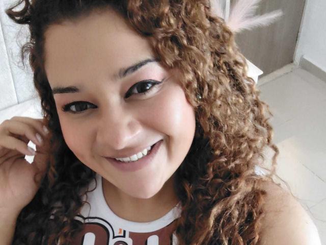 Close-up photo of NataliaWendys with curly brown hair, smiling at the camera. She has medium brown skin, dark eyebrows, and black eyeliner. Her white teeth are visible, and she's wearing a sleeveless white shirt with red and black text. Her right hand is touching her hair. The image has natural light, highlighting her facial features. The background is blurred, focusing on NataliaWendys' face. She has a confident, friendly expression.