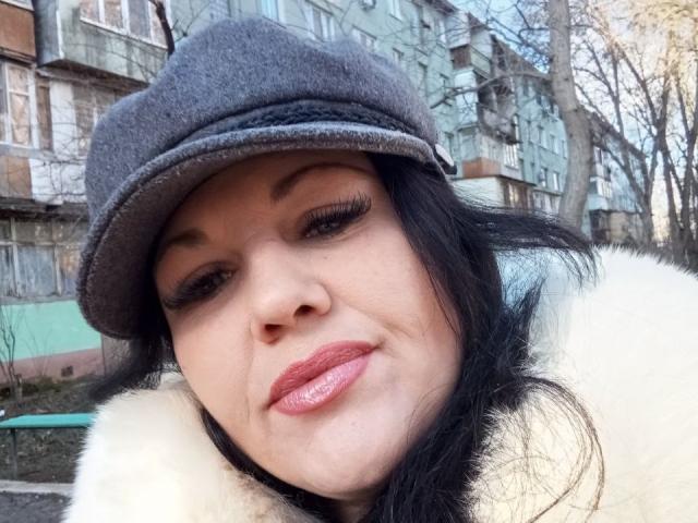 ZoieParker with grey flat cap, black hair, and shiny pink lipstick, smiling slightly, wearing a white fur coat, standing outdoors with a gray building in the background.