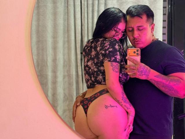 JackAndTriana with long black hair, floral top, and thong showing her big ass, standing next to tattooed Jack holding a phone, in a mirror selfie. Both have tattoos.
