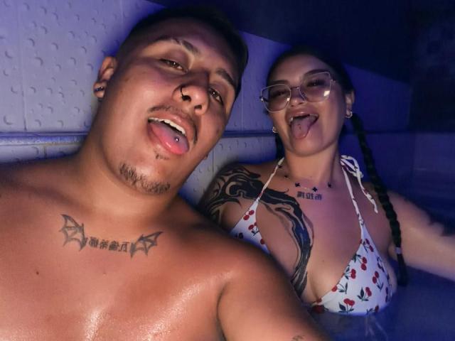 JackAndTriana with tattoos, both sticking out tongues, in a hot tub. Jack's shirtless with a nose ring, Triana has glasses, braids, and a cherry bikini top.
