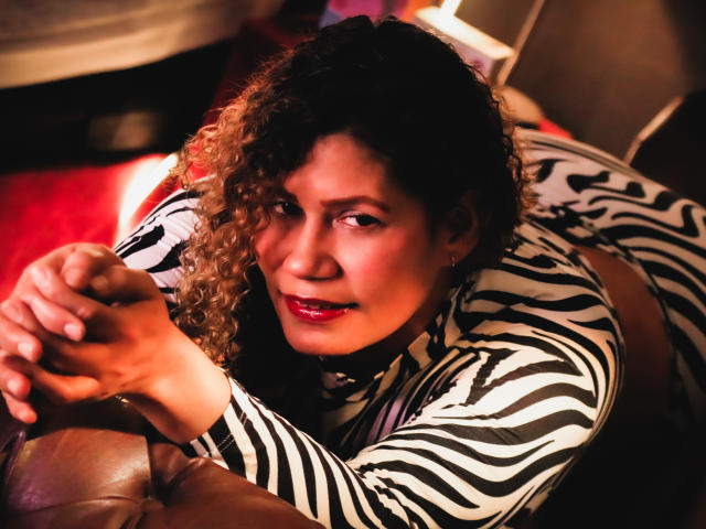 Zoe Weber, con i capelli castano ricci, il rossetto rosso e una maglietta a righe zebrate, sdraiata su un divano in pelle marrone, con le mani vicino al viso, che guarda la fotocamera con un leggero sorriso..