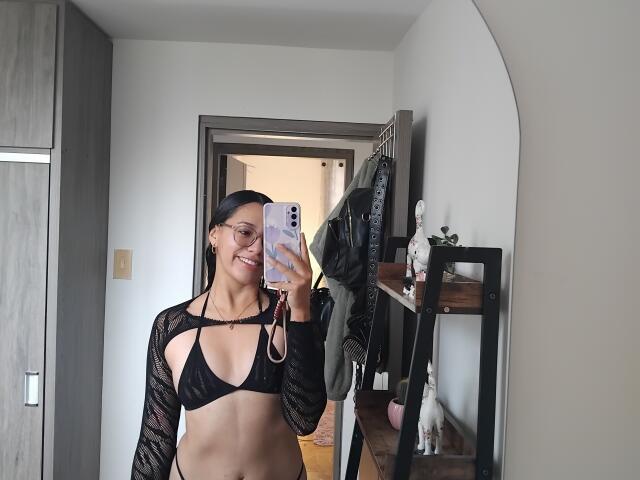 Mllevv with black bikini top and long-sleeve mesh shirt, showing tits and smiling at mirror selfie, holding pink phone, in bedroom with door and clothes rack.