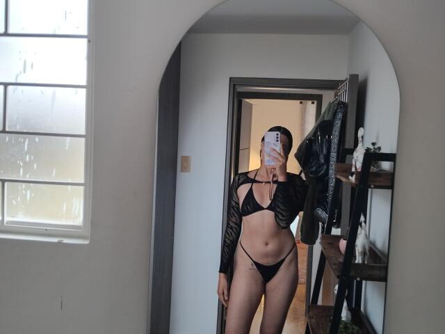 Mllevv trägt schwarze, durchsichtige Dessous und zeigt ihre Brüste in einem String. Sie steht vor einem Spiegel, wobei ein Handy ihr Gesicht verdeckt. Rechts daneben befindet sich ein Holzregal mit Kleidung..