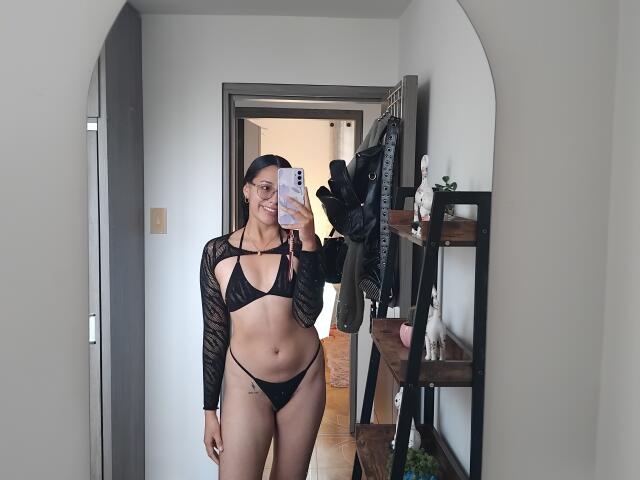 Vrouw met zwarte kanten mouwen, zwart string bikini, en een kleine tatoeage aan de linkerkant, die een telefoon vasthoudt, lachend, staand in een gang, spiegelbeeld, zichtbaar schaamtegebied, slank, middelgrote borsten..