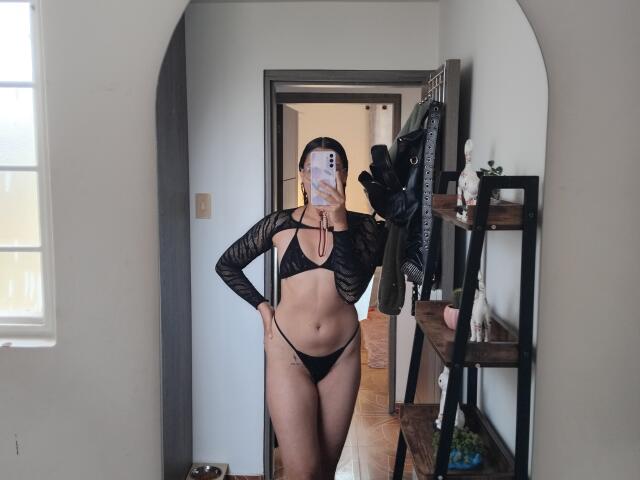 Mllevv draagt een zwarte top van mesh en een string, waarbij haar platte buik en kleine billen zichtbaar zijn. Ze staat in een gang en maakt een selfie in de spiegel met haar telefoon..
