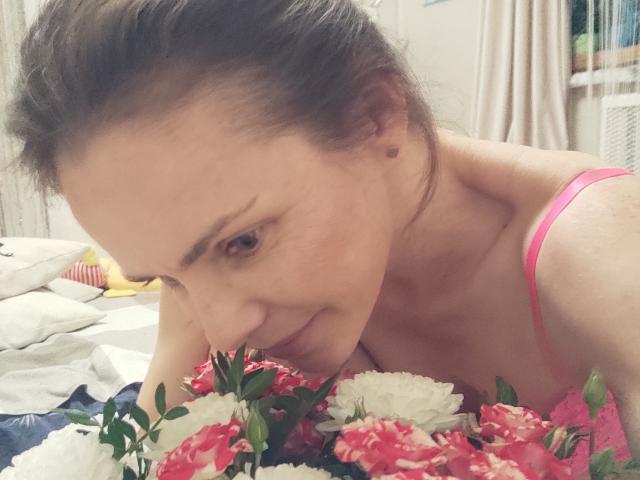 SeaDream with brown hair, pink strap, and fair skin, leans over colorful flowers, nose close, smiling softly. Boobs partially visible, pink strap over shoulder. Bedroom setting.
