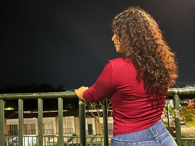 NicollWest with curly brown hair, red long-sleeve shirt, and blue jeans, leaning on a green railing at night, showing her back and butt.