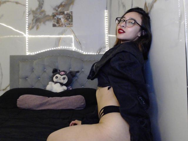 CamiVargas con gli occhiali, rossetto rosso, maglietta nera e biancheria intima nera, appoggiata al muro, che mostra il suo sedere scoperto, seduta sul letto con un gatto di peluche e un cuscino viola..
