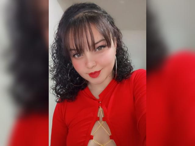 GiaDonovan, com cabelo preto encaracolado, batom vermelho e um vestido vermelho com um detalhe dourado, está olhando para o reflexo no espelho, mostrando seus seios. Ela está sorrindo e tem um piercing no nariz..