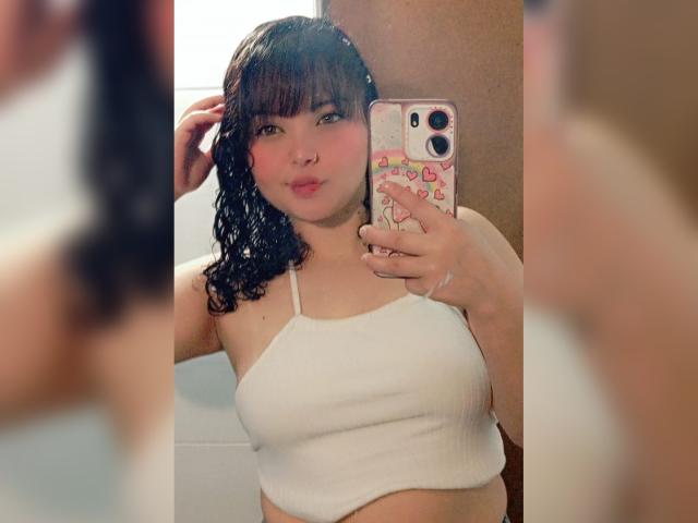 GiaDonovan, avec ses cheveux noirs mouillés, ses lèvres roses et pulpeuses, portant un crop top blanc côtelé, tenant un téléphone rose avec des autocollants en forme de cœur, prenant un selfie devant un miroir. Seins visibles..