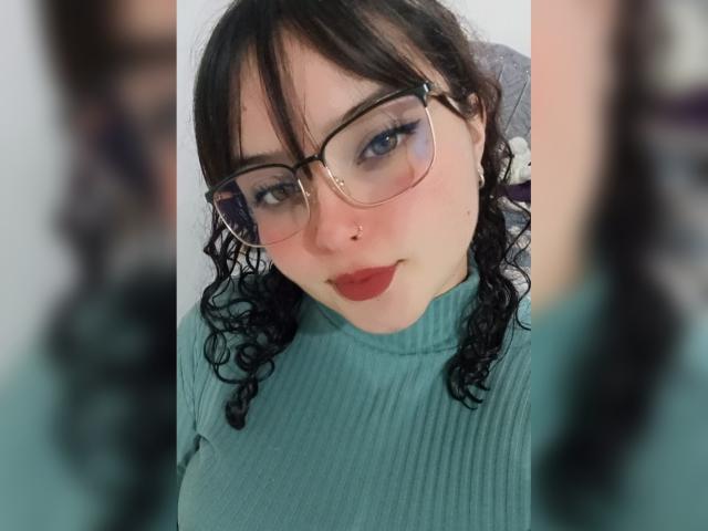 GiaDonovan tem cabelo preto encaracolado, usa óculos e batom vermelho. Ela está vestindo um suéter verde canelado, olhando para baixo com um leve sorriso. Seus seios são de tamanho médio e visíveis..