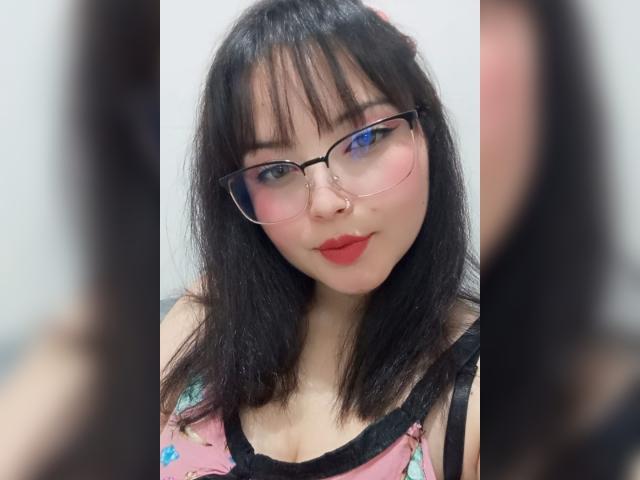 GiaDonovan, con i capelli neri, gli occhiali, il rossetto rosso, il piercing al naso e una maglietta rosa con fiori, che mostra il seno in un selfie. Riflessi sfocati nello specchio incorniciano il suo viso..