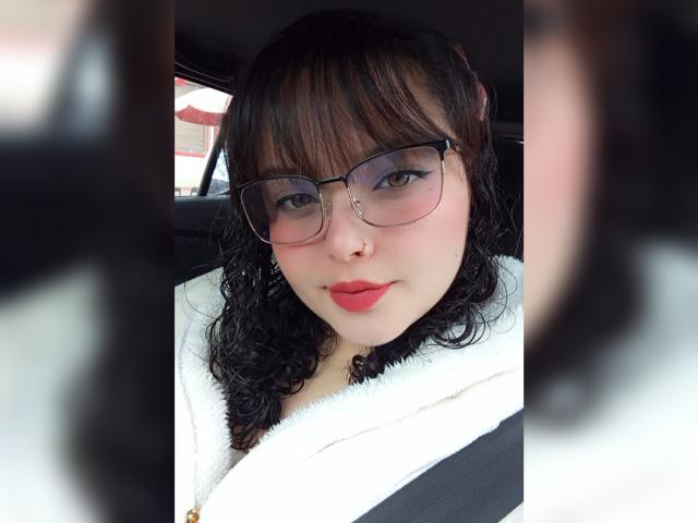 GiaDonovan, con cabello negro rizado, gafas, lápiz labial rojo, abrigo blanco esponjoso y cinturón de seguridad, mirando a la cámara, mostrando un rostro lindo y atractivo, con un ligero rubor en las mejillas y un piercing en la nariz..