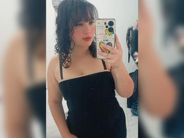 GiaDonovan, con i capelli neri ricci, indossa un abito nero senza spalline, sta scattando un selfie allo specchio, mostrando il seno, tiene in mano un telefono con adesivi colorati, in una stanza bianca..
