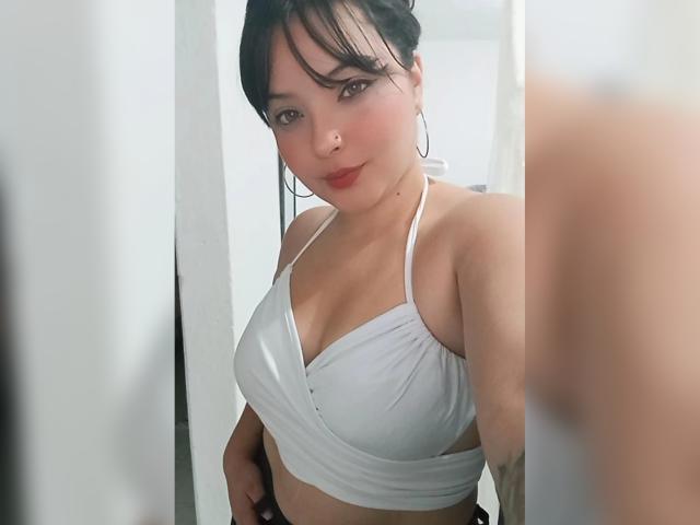 GiaDonovan mit mittelgroßer Brust, trägt ein weißes Top mit Halter, hat dunkles, zu einem unordentlichen Dutt gebundenes Haar, große Ohrringe und ein leichtes Lächeln und steht in einem Spiegel-Selfie..