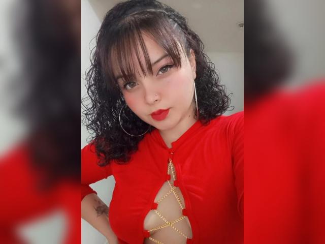 GiaDonovan with curly black hair, red lipstick, and hoop earrings, wearing a red top with a gold chain cutout, showing her small tits and tattoo on her right arm.
