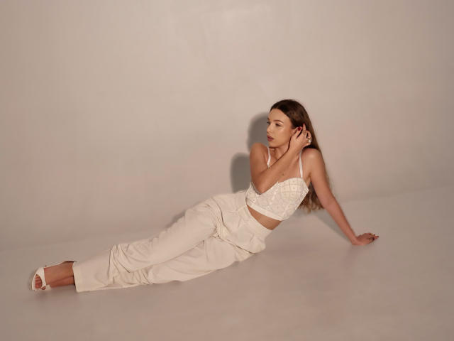 LilyBlake is lying down, wearing a white lace bralette and matching high-waisted pants, with her hand on her head, showing her tits and ass. She's wearing white heels.