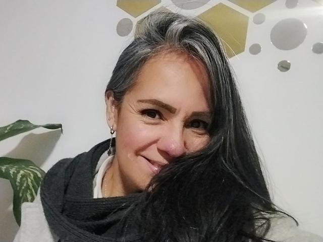 LailaCastillo with black hair, smiling, wearing a grey scarf, white shirt, and earrings, her tits partially visible, green plant on the left, white wall background.