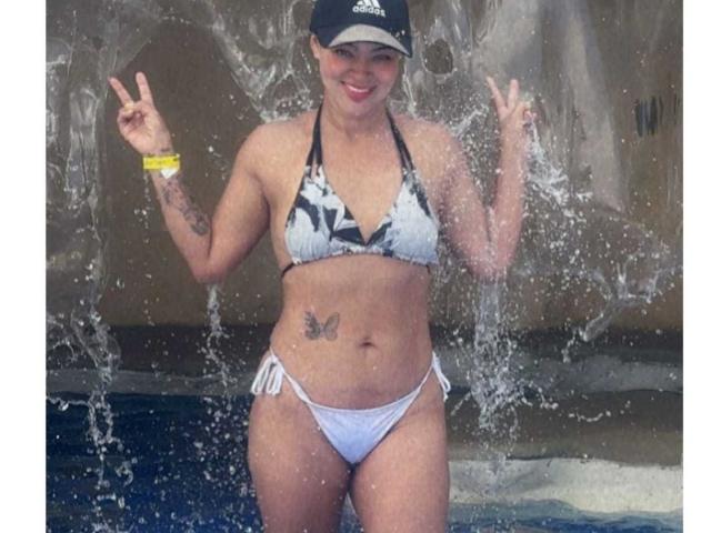 AlyssiaOcean stands in water, flashing dual peace signs, wearing a white and black patterned bikini, black cap, yellow bracelet, butterfly tattoo on her belly, and smiling.