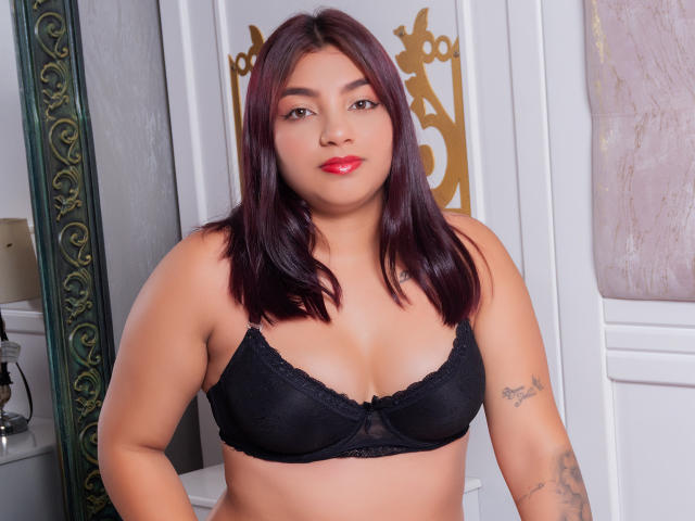SamanthaSimon with medium brown skin, red lipstick, and straight dark red hair, wearing a black lace bra, showing her tits and tattoo on her right arm, smiling.